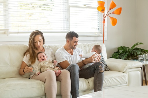 A beautiful family portrait of a new family of hispanic descent holding dizygotic twin boys. The attractive couple are playing with their babies on the couch and smiling.