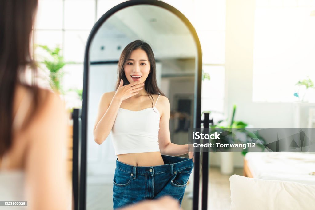 young woman wearing large-size jeans , looks at her figure in front of the mirror, very happy that she has succeeded in losing weight. Weight Stock Photo