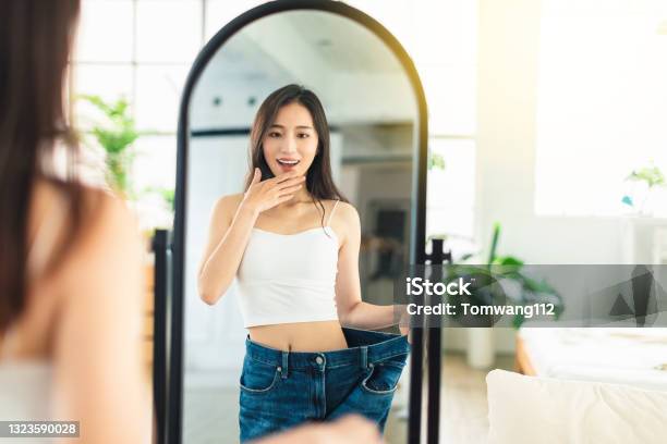Young Woman Wearing Largesize Jeans Looks At Her Figure In Front Of The Mirror Very Happy That She Has Succeeded In Losing Weight Stock Photo - Download Image Now