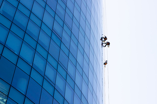 Window washers hanging outside blue glass modern building. Risky job, dangerous work concepts