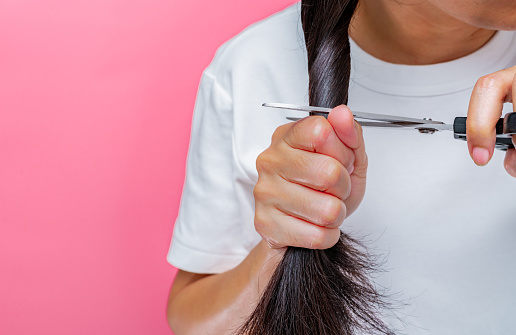Women cutting damaged and dry hair. Asian woman cutting hair with scissors for donating to cancer patients. Hair donation for breast cancer person. Woman with black long hair on pink background.