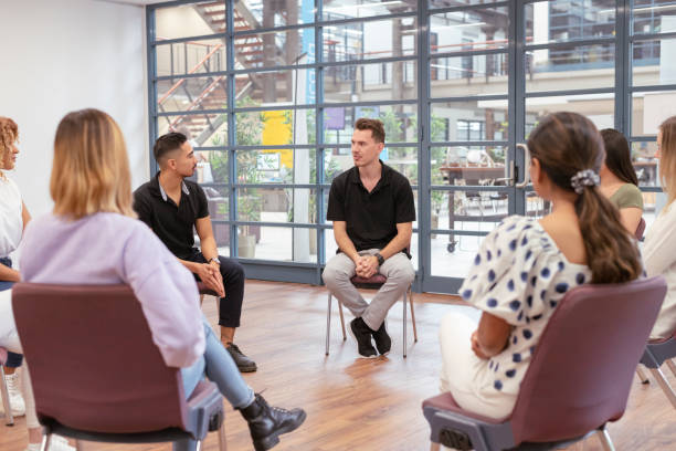 Multi ethnic group of young adults in meeting A young man shares his emotions and experiences with a supportive mixed-race group of people while attending group therapy. Vulnerability, community and mental health concepts. university students australia stock pictures, royalty-free photos & images