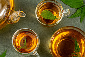 Cups of herbal tea, transparent teapot and nettle leaves on green background. Calming drink concept. Top view Flat lay