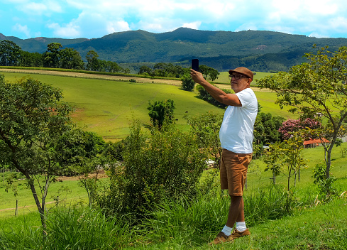 Retired old man standing in a beautiful green field taking a selfie around a beautiful mountain landscape.