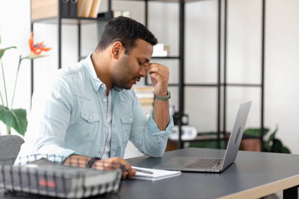 tired indian businessman having headache or migraine sitting at the office desk, young man freelancer sitting at home office suffering from overwork, feel eye strain from computer - human eye tired rubbing businessman imagens e fotografias de stock