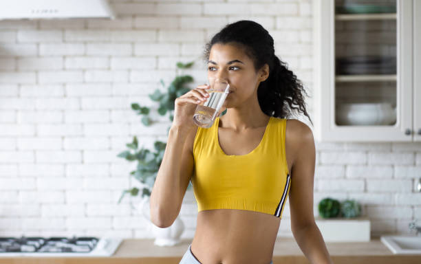 young african american woman with beautiful body drinks fresh clean water after fitness workout standing in the home kitchen healthy lifestyle - drinking water stockfoto's en -beelden