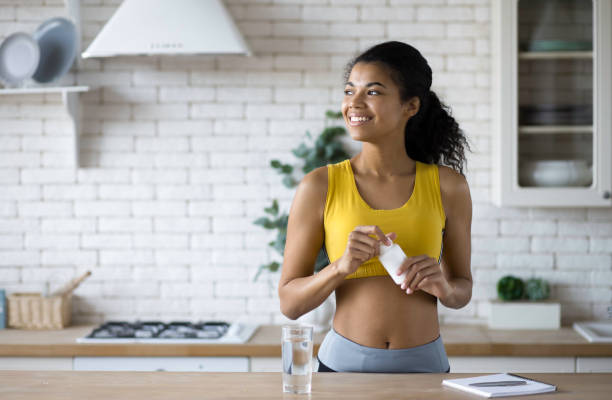 feliz mujer deportiva afroamericana en ropa deportiva sosteniendo una botella de suplementos nutricionales, mirando felizmente por la ventana, estilo de vida saludable - vitamin pill fotografías e imágenes de stock