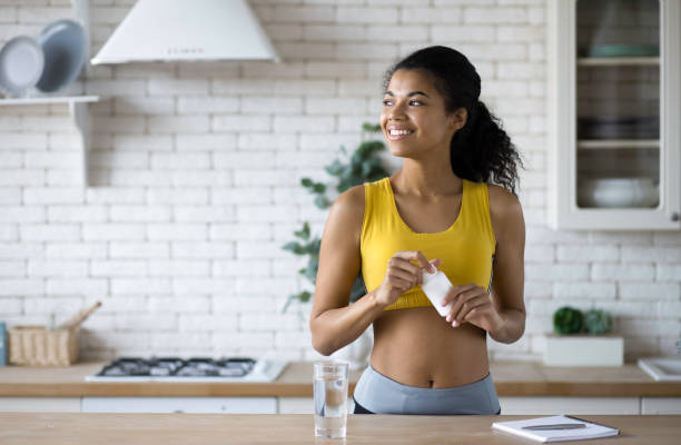 스포츠웨어에 있는 행복한 스포츠아프리카계 흑인 여성이 영양 보충제 한 병을 들고 창 밖으로 행복하게 바라보며 건강한 라이프스타일을 보여줍니다. - nutritional supplement 이미지 뉴스 사진 이미지