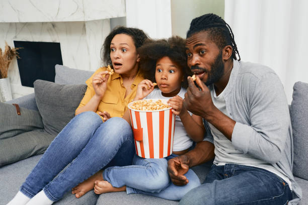 padre, madre e figlio siedono sul divano con i volti spaventati durante la visione di film spaventosi a casa - home movie foto e immagini stock