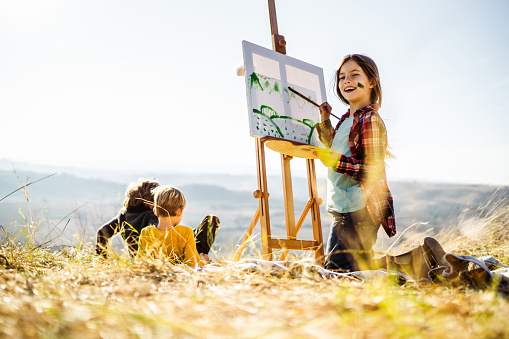 Happy little girl making a painting of her friends during autumn day on a hill and looking at camera. Copy space.