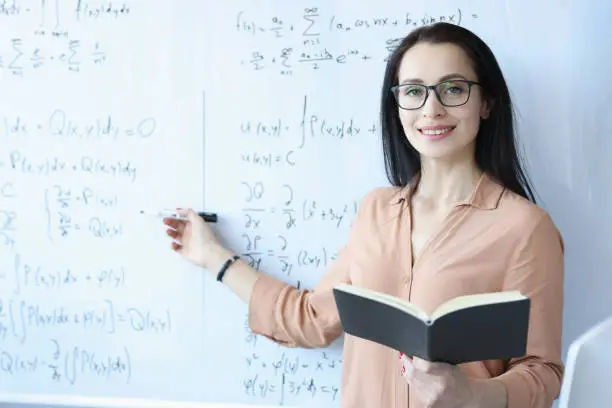 Woman mathematician with glasses standing at blackboard with formulas and holding open book. School education concept