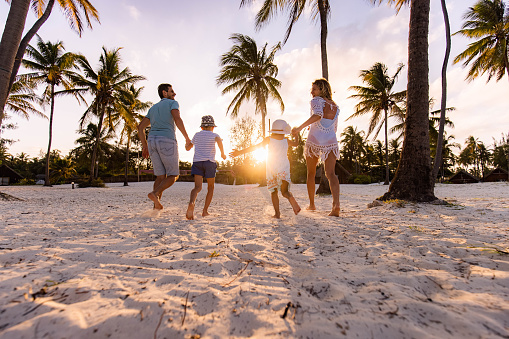 Rear view of happy parents and their small sons having fun while holding hands and running on the beach at sunset. Copy space.