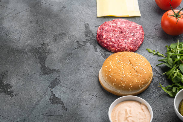 Ingredients for cooking burgers. Minced beef patties, buns, tomatoes, herbs and spices, on gray stone background, with copy space for text