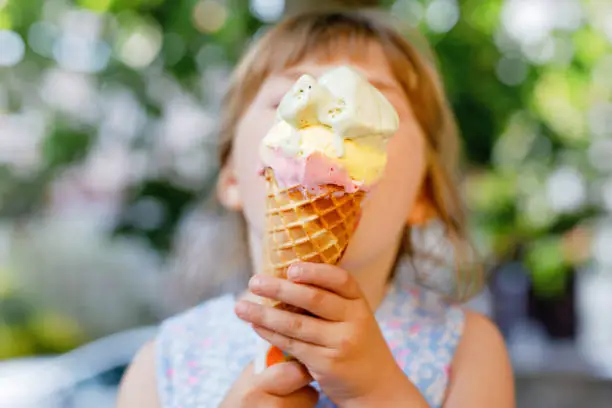 Little preschool girl eating ice cream in waffle cone on sunny summer day. Happy toddler child eat icecream dessert. Sweet food on hot warm summertime days. Bright light, colorful ice-cream.