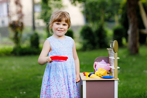 Little preschool girl playing with toy kitchen in garden. Happy toddler child having fun with role activity game preparing food. Children play outdoors in summer