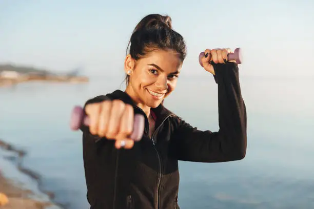 Cheerful young sportsgirl doing exercises with small dumbbells at the seaside in the morning