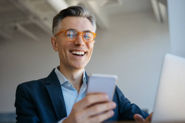 reife mann mit laptop-computer, halten handy sitzen in modernen büro, erfolgreiches geschäft. porträt von schönen emotionalen freiberuflern erhalten zahlung, arbeiten von zu hause aus - smart phone business person computer businessman stock-fotos und bilder