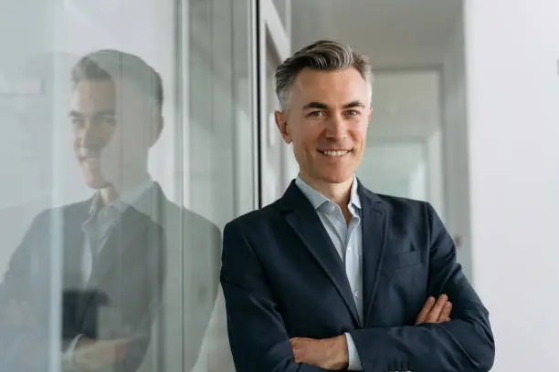 Photo of Handsome mature manager with arms crossed looking at camera, smiling standing in modern office