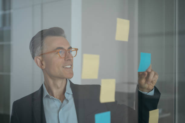 istock Mature scrum master wearing stylish eyeglasses using sticky notes standing near planning board in modern office. Agile methodology, scrum concept 1323649987