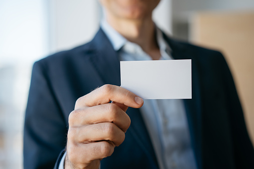 Close up of man holding blank business card with copy space, standing in modern office. Mock up, advertisement concept