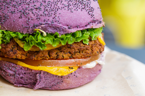 Close up color image depicting freshly prepared vegan burgers - loaded with salad and sauce, and sandwiched inside a brightly-colored bun topped with seeds.