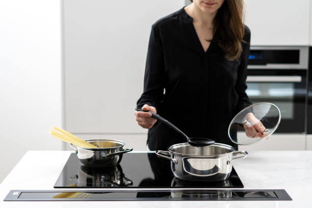 Woman cooking spaghetti in modern white kitchen Cropped shot of woman boiling water in metal pot to cook spaghetti, holding glass lid and big spoon while cooking on black ceramic induction cooktop in white minimalistic kitchen large pasta pot stock pictures, royalty-free photos & images