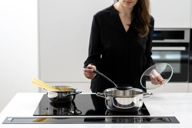 Woman cooking spaghetti in modern white kitchen Cropped shot of woman boiling water in metal pot to cook spaghetti, holding glass lid and big spoon while cooking on black ceramic induction cooktop in white minimalistic kitchen glass ceramic stove top stock pictures, royalty-free photos & images