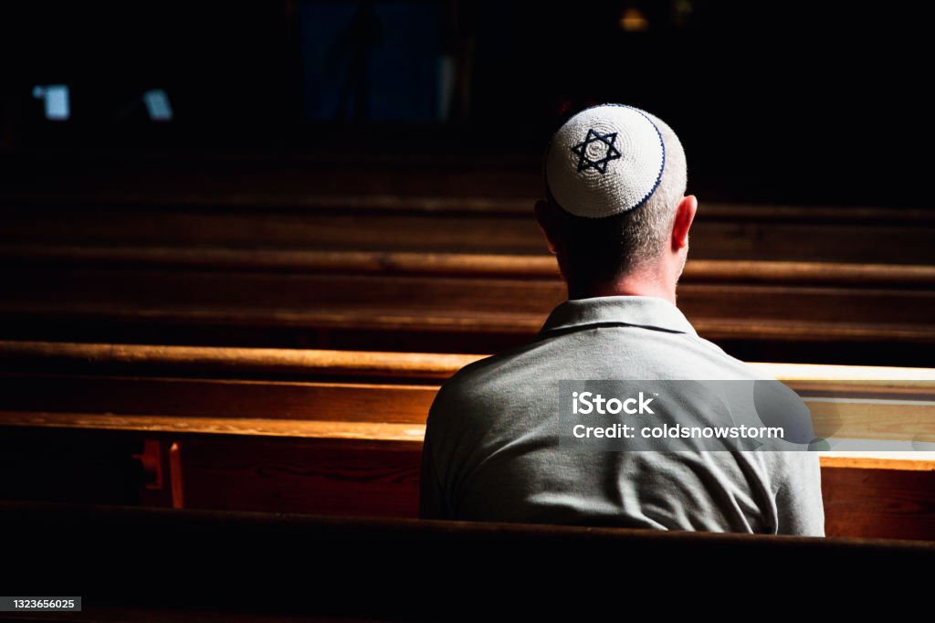 Young Jewish man wearing skull cap praying inside synagogue Close up image depicting a young caucasian Jewish adult man in his 30s inside a synagogue. He has his head bowed in prayer and he is wearing the traditional Jewish skull cap - otherwise known as a kippah or yarmulke - on his head. The man has a beard and the background of the synagogue is blurred out of focus. Horizontal color image with copy space. Judaism Stock Photo