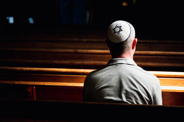 Young Jewish man wearing skull cap praying inside synagogue Close up image depicting a young caucasian Jewish adult man in his 30s inside a synagogue. He has his head bowed in prayer and he is wearing the traditional Jewish skull cap - otherwise known as a kippah or yarmulke - on his head. The man has a beard and the background of the synagogue is blurred out of focus. Horizontal color image with copy space. synagogue stock pictures, royalty-free photos & images