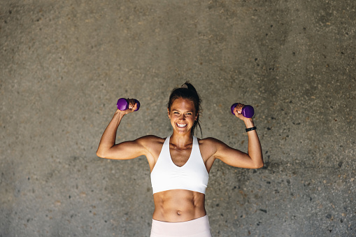 Smiling woman exercising with hand weights. Woman holding dumbbell flexing her biceps looking at camera and smiling against a wall.