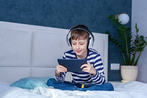 Cute child boy in headphone playing on a digital tablet on the bed in living room. Education, free time, technology and internet concept
