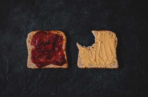 Strawberry jam and peanut butter sandwich/toast. PBJ/Peanut butter and jelly spread. Black stone background, bite taken. Smooth colors, nice rye bread.