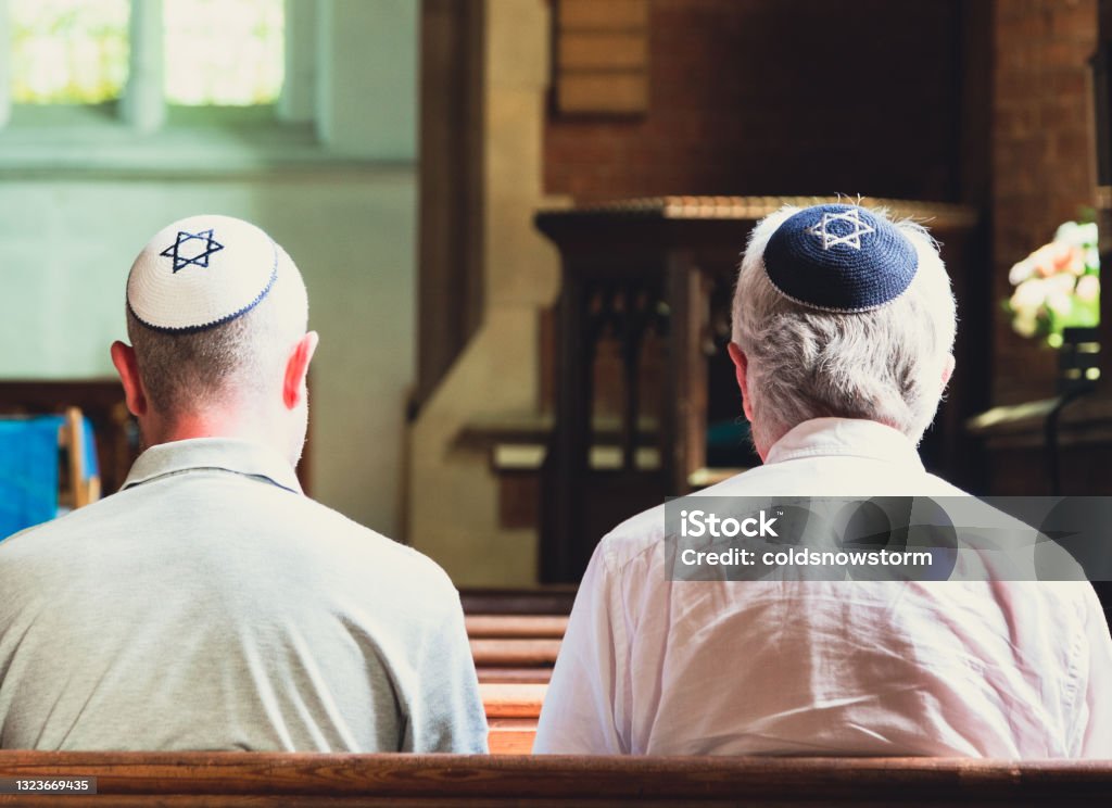 Jewish men sitting together and praying inside synagogue Close up image depicting a rear view of two Jewish men sitting together inside a synagogue. They have their heads bowed in prayer and they are wearing the traditional Jewish skull cap - otherwise known as a kippah or yarmulke - on their heads. Horizontal color image with copy space. Rabbi Stock Photo