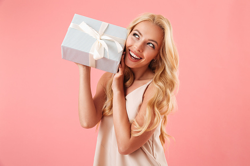 Portrait of a smiling young woman holding present box at her ear isolated over pink background