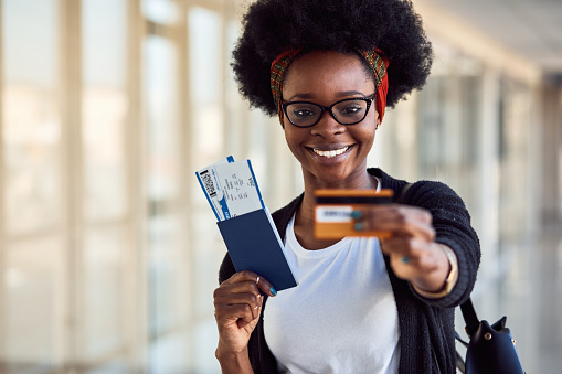 Young african american female passanger in casual clothes is in airport holding tickets and credit card.