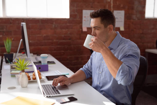 istock Caucasian businessman sitting at desk in office using laptop and drinking coffee 1323674112