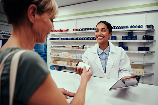 Young happy female pharmacist giving bottle of medicines to customer while holding digital tablet