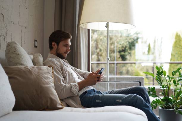 istock Handsome man in basic t-shirt smiling and holding mobile phone in hands while sitting on couch in living room. 1323678455