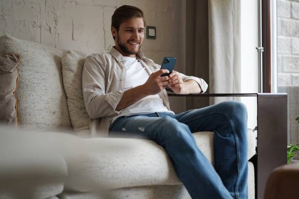 istock Handsome man in basic t-shirt smiling and holding mobile phone in hands while sitting on couch in living room. 1323678461