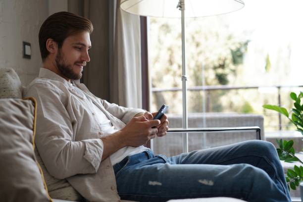 istock Handsome man in basic t-shirt smiling and holding mobile phone in hands while sitting on couch in living room. 1323678620