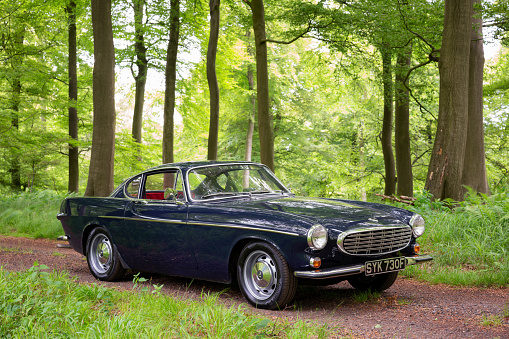 Stokenchurch, UK - June 11, 2021: A classic dark blue Volvo 1800S from 1967 sitting on a track running through beautiful woodland in the English countryside. Made famous when a white version was used by Roger Moore in the iconic sixties tv show The Saint, this svelt Swedish sports coupe is still a head-turner to this day.