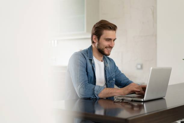 istock Young casual man working on laptop online, sitting at table in kitchen, looking at computer screen 1323680061