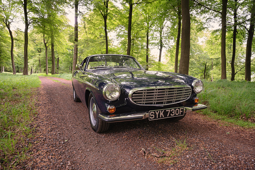Stokenchurch, UK - June 11, 2021: A classic dark blue Volvo 1800S from 1967 sitting on a track running through beautiful woodland in the English countryside. Made famous when a white version was used by Roger Moore in the iconic sixties tv show The Saint, this svelt Swedish sports coupe is still a head-turner to this day.