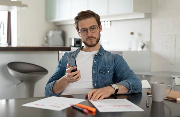 istock Confident man sitting at a table holding smartphone, chatting online with colleagues, looking at documents 1323681248