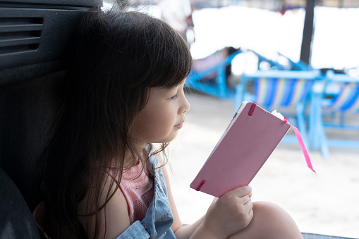 cute little girl sitting in the back of a car reading a pink book near the beach. weekend vacation