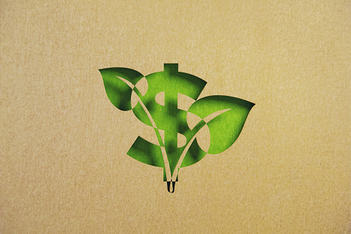 Cut out leaf shaped made of recycled paper intersect with American dollar sign on green background. Horizontal composition with copy space. Sustainability concept.