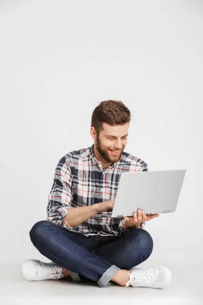 Portrait of a smiling young man in plaid shirt sitting on a floor with laptop computer isolated over white background
