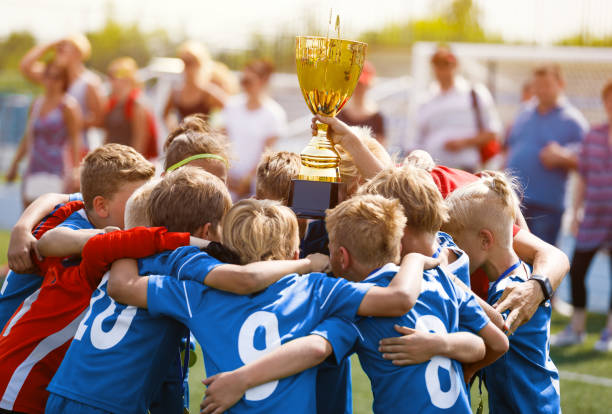 niños felices en el equipo de fútbol ganando la copa de oro en el torneo escolar. young boys in blue soccer uniforms rising up golden cup. joven entrenador celebrando el éxito con los jugadores de fútbol infantil - winning achievement award little boys fotografías e imágenes de stock