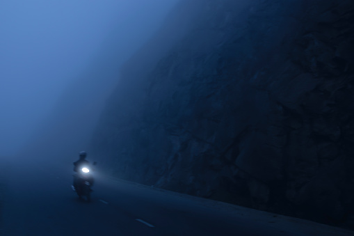 Motorcycle driver riding on the misty mountain road in Sa Pa, North Vietnam. Motorbike with light trail from headlight leading through the dark misty. Exploration, adventure, travel concepts. Blur motion.
