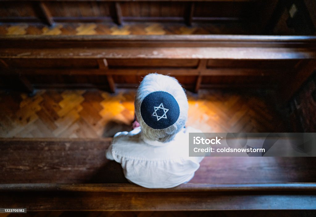 Senior Jewish man wearing skull cap praying inside synagogue Close up image depicting a senior caucasian Jewish adult man in his 60s sitting inside a synagogue. He has his head bowed in prayer and he is wearing the traditional Jewish skull cap - otherwise known as a kippah or yarmulke - on his head. Horizontal color image with copy space. Synagogue Stock Photo